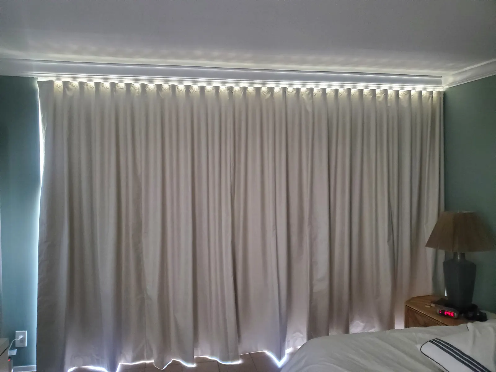 Ivory blackout-lined draperies with a warm LED light strip along the crown molding, framing a bedroom window beside a wood table lamp