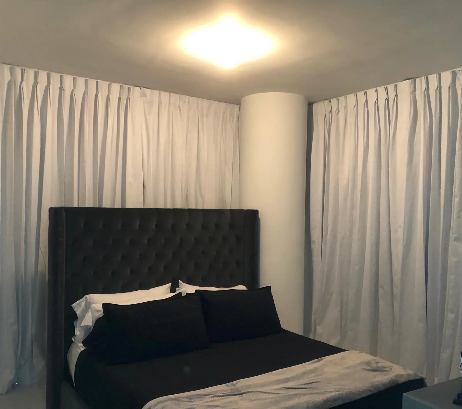 Champagne pinch-pleat draperies wrapping a curved bedroom corner behind a tufted upholstered headboard