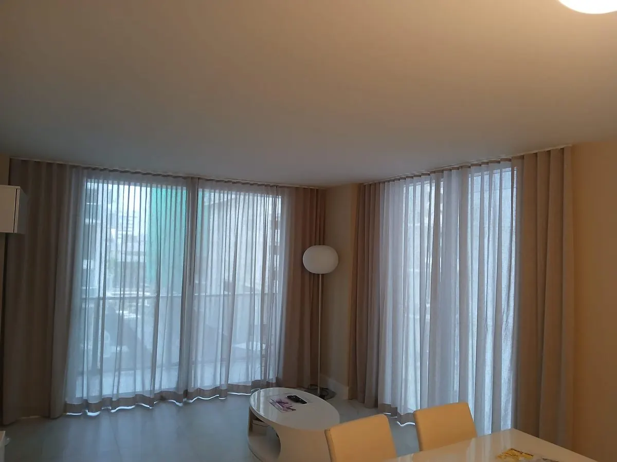 Sheer pinch-pleat draperies paired with taupe side panels across corner floor-to-ceiling windows in a living and dining space, daylight streaming through