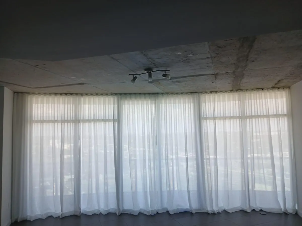 Floor-to-ceiling white ripplefold sheer draperies drawn along a continuous wave-fold track beneath an exposed concrete ceiling