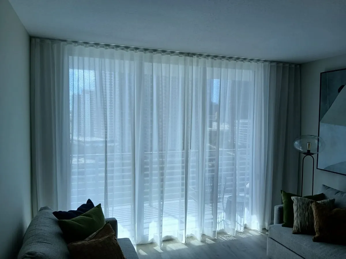 Sheer white draperies filtering evening light across a living room window, gray sofas with green and brown accent pillows in the foreground