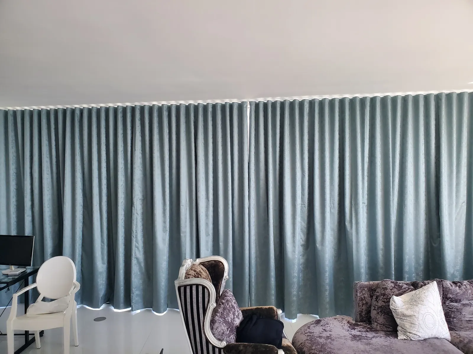 Sage-blue wave-fold draperies framing floor-to-ceiling windows in a high-rise living room with a striped wingback chair and crushed-velvet sofa