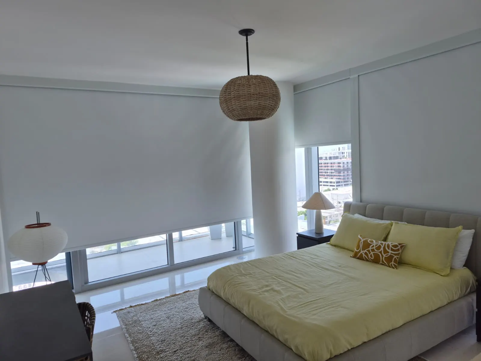 A blackout roller shade lowered over a bedroom window, light blocked over a yellow-upholstered bed with the city skyline visible through the uncovered strip below