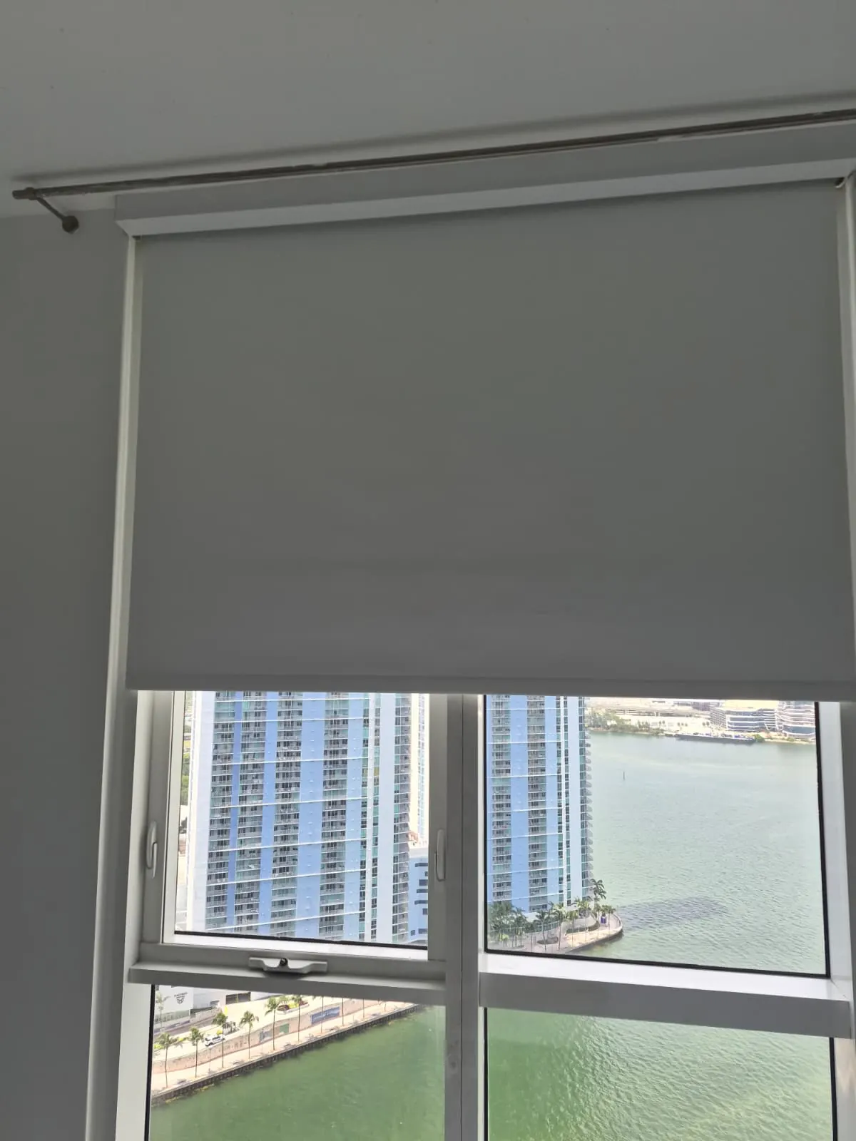 Grey blackout roller shade partially lowered over a high-rise window, with blue waterfront towers and Biscayne Bay visible below
