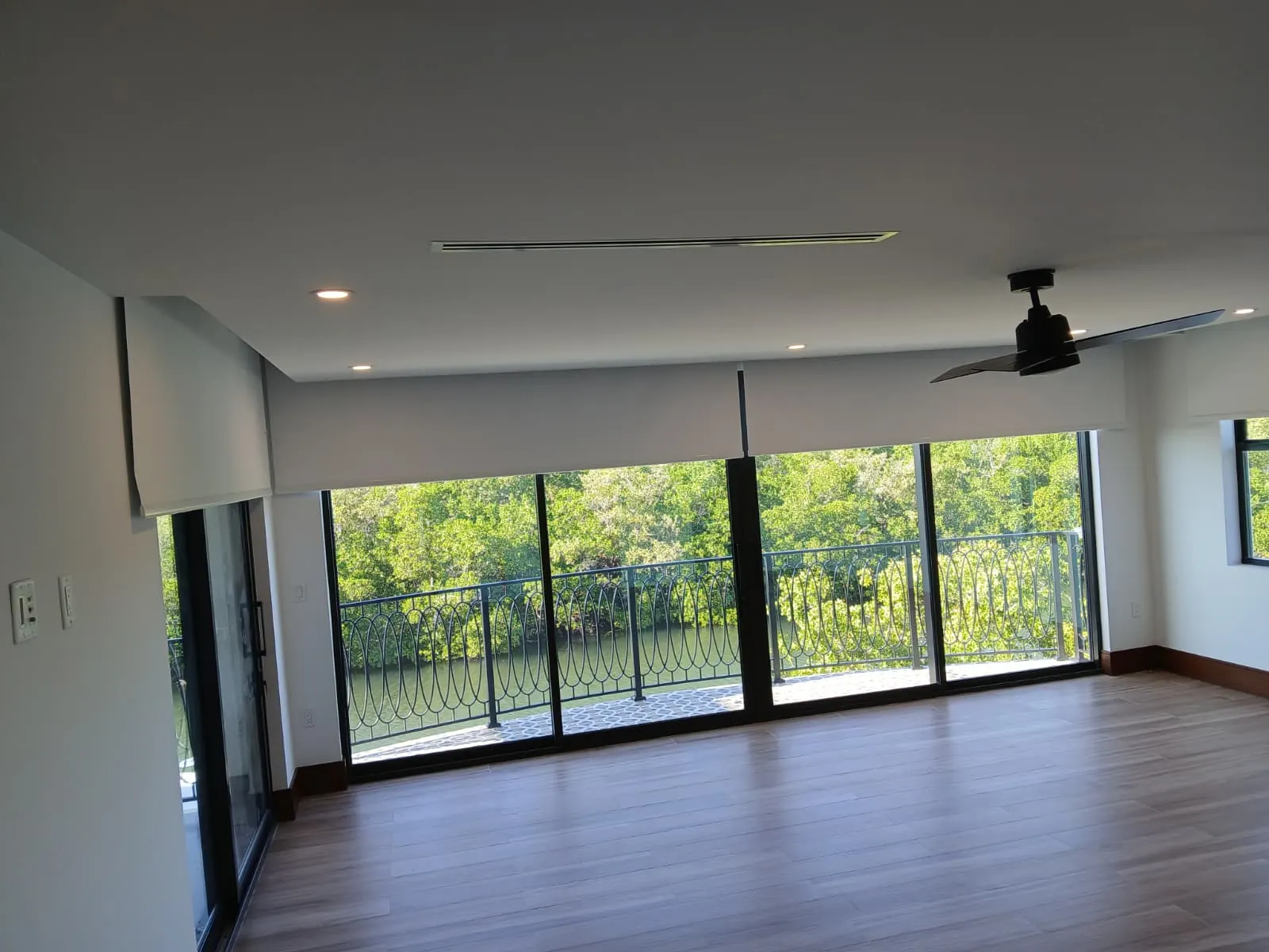 White blackout roller shade with valance partially lowered in an open living space, overlooking a wrought-iron balcony railing and a lush mangrove river