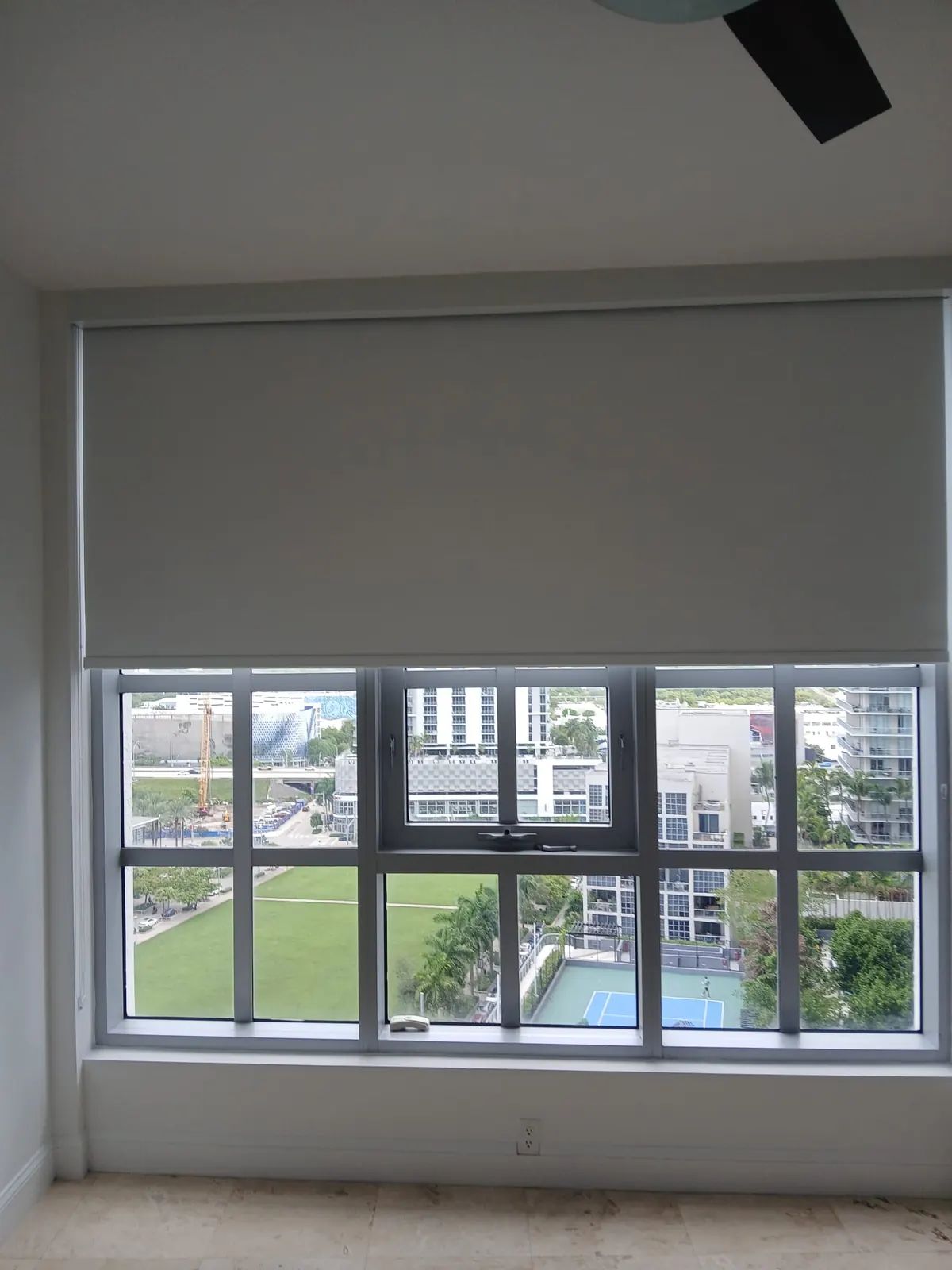 Grey blackout roller shade partially lowered over a wide window, overlooking a landscaped park, tennis court, and palm trees below