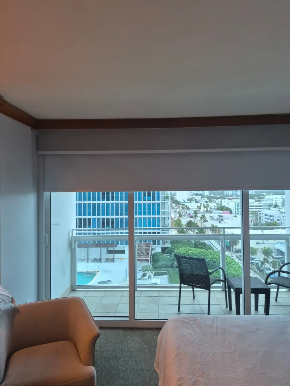 Grey blackout roller shade lowered over a balcony sliding door, pool deck and palm-lined street visible below