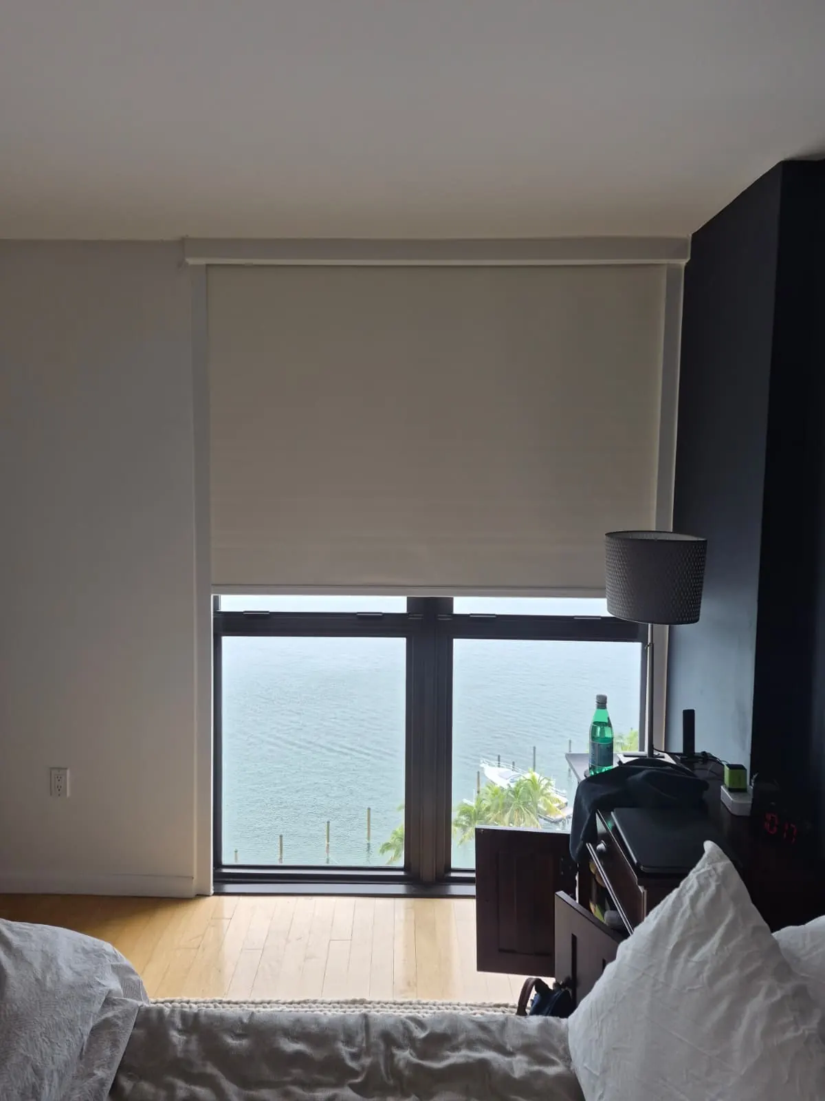 Cream blackout roller shade partially lowered in a bedroom, overlooking open water, palm trees, and docked boats