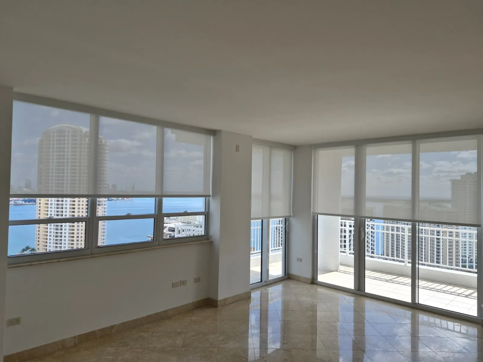 Solar screen roller shades lowered over a corner window wall in an empty high-rise living room, sheer fabric revealing a bay and marina view beyond