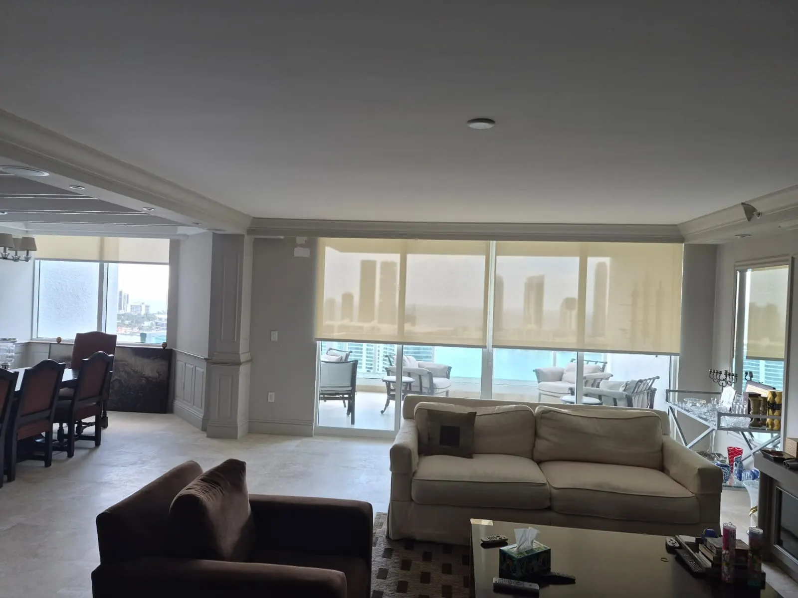 Sheer solar screen roller shades filtering daylight across a living and dining room's floor-to-ceiling windows, city skyline and water visible through the fabric