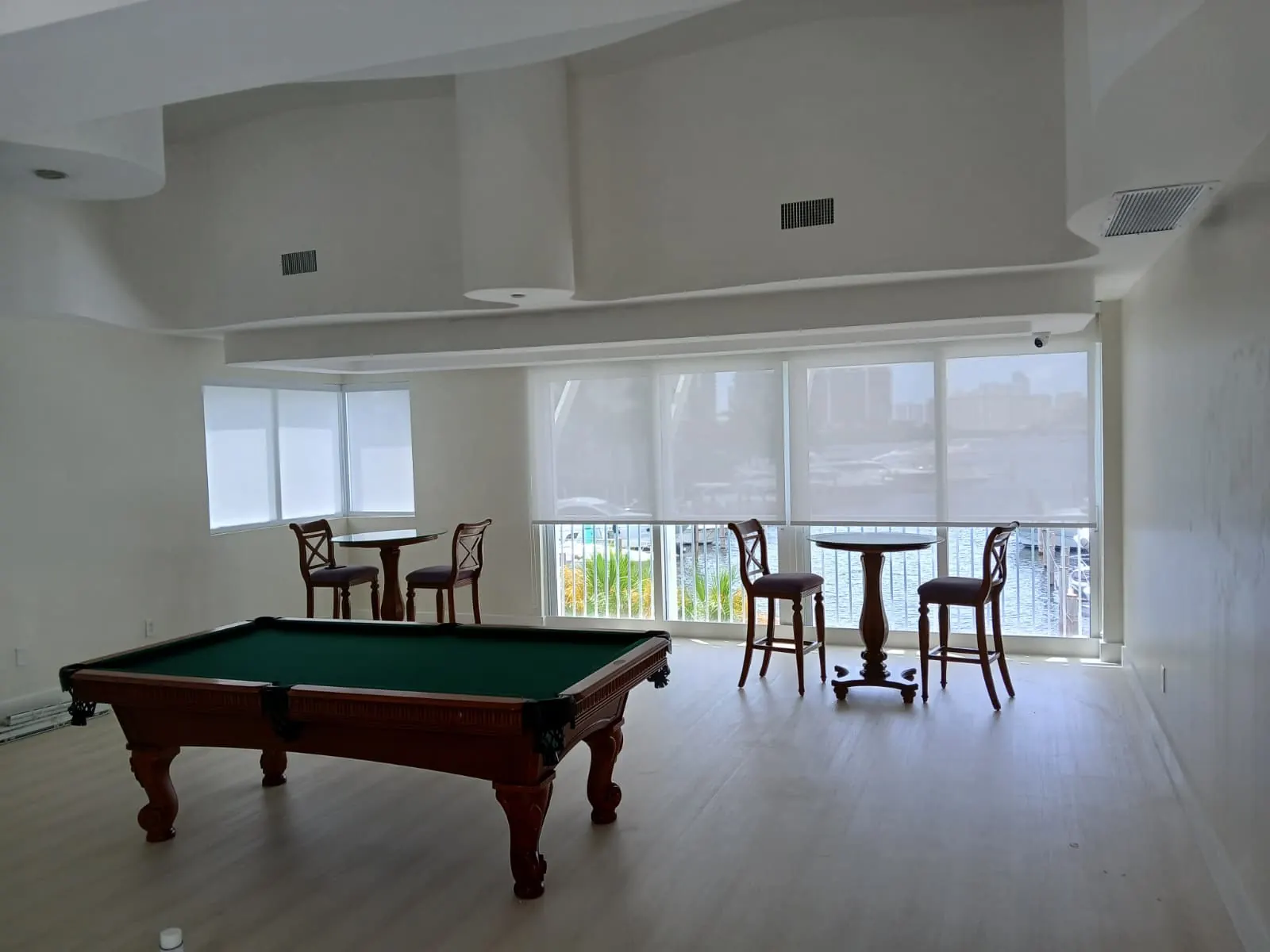 Solar screen roller shades lowered across floor-to-ceiling windows in a marina-front game room, translucent fabric revealing docked boats and the waterway beyond