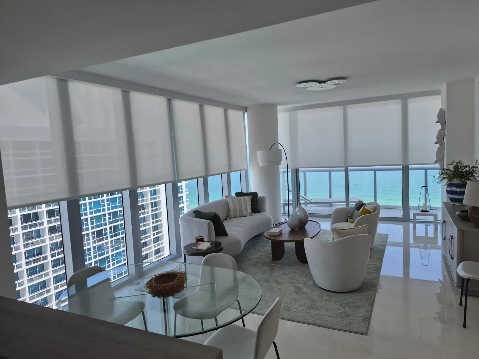 Solar screen roller shades lowered across a corner living and dining room, sheer white fabric filtering bright daylight over a turquoise ocean view