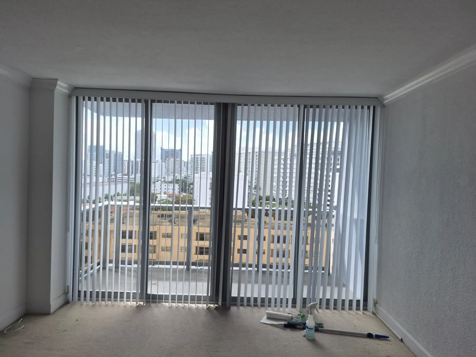 White vertical blinds hung across a wide corner window wall, opening onto a panoramic city skyline