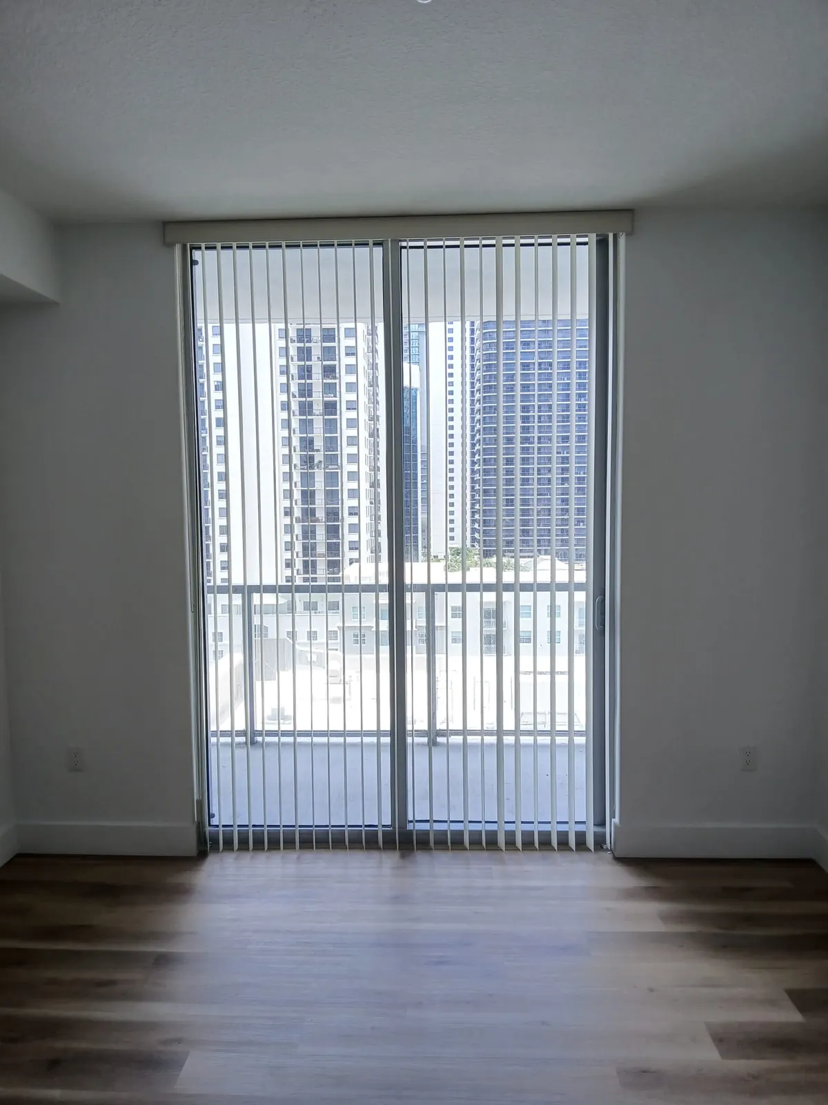White PVC vertical blinds hanging straight across twin sliding glass doors, framing a downtown high-rise skyline beyond