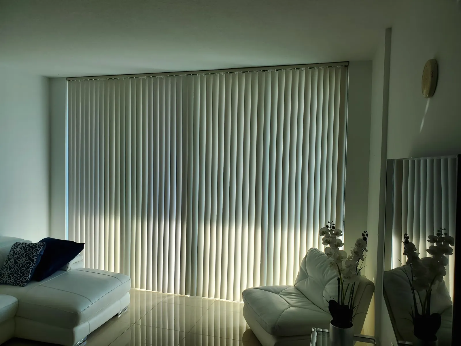 Cream fabric vertical blinds diffusing warm evening light across a modern living room with white leather seating