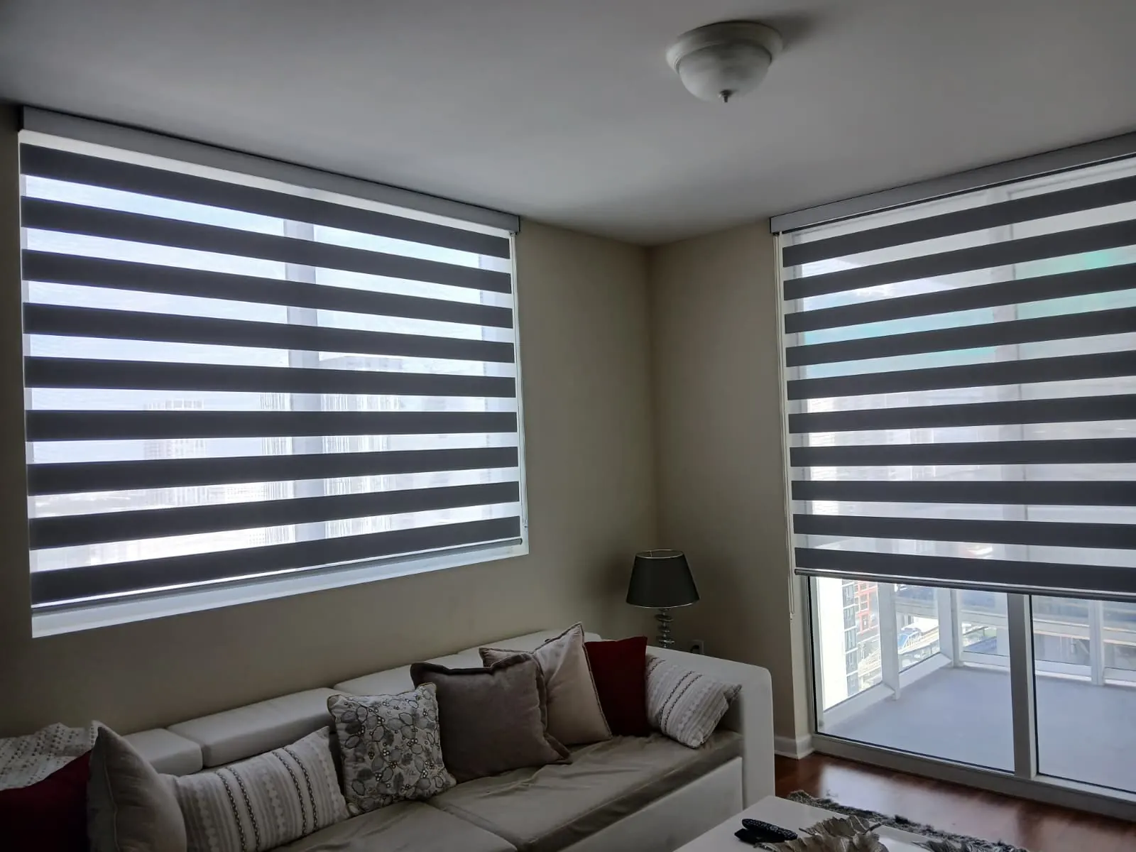 Two zebra shade panels in charcoal and white stripes filling a pair of living room windows, with the city skyline visible through the sheer bands above a plush sectional sofa