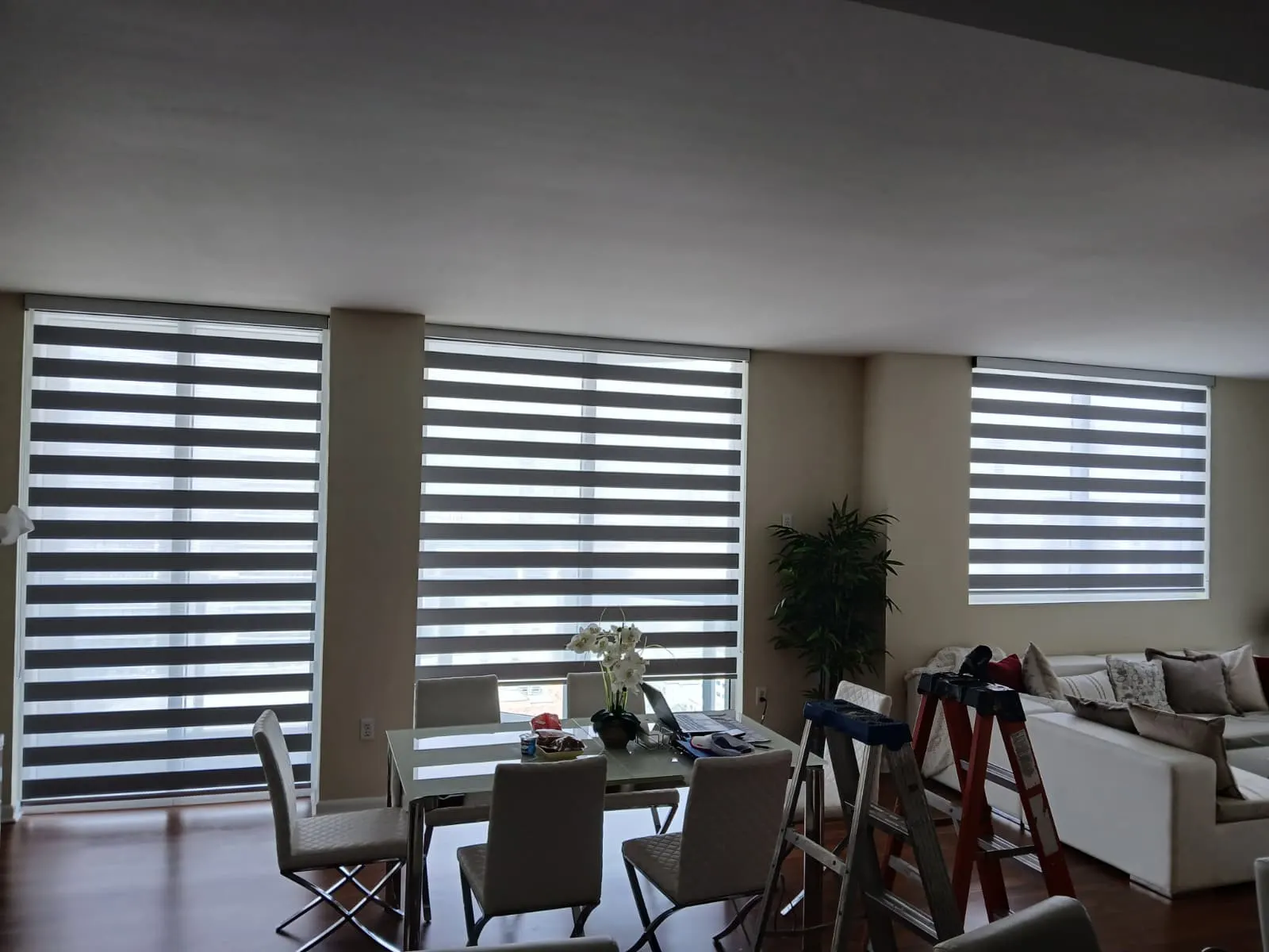 Three zebra shades in charcoal and white stripes spanning a living and dining room's windows, a dining table in the foreground